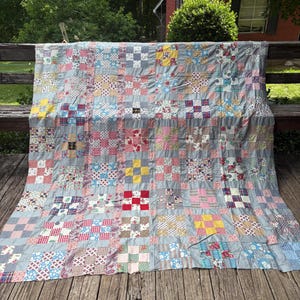 May include: A patchwork quilt featuring a grid of square blocks in various floral, plaid, and gingham patterns. The blocks are separated by solid light gray sashing strips, creating a repeating geometric design with a mix of red, blue, yellow, and white colors.