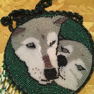 May include: A circular beaded pendant with a wolf design. The pendant features a white and grey wolf face with black eyes and nose, framed by a green beaded border. White and green beaded fringe hangs from the base.