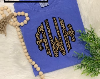 May include: A light blue t-shirt with a leopard print monogram "AWK" in a circle. The shirt is on a white fluffy surface.
