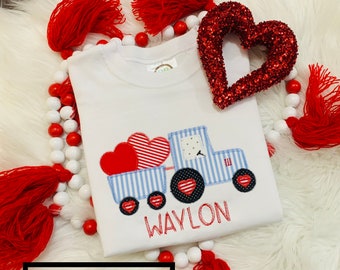 May include: White toddler t-shirt with a red, blue, and white embroidered design of a truck carrying hearts. The truck has the name "WAYLON" embroidered below it. The shirt is surrounded by red and white beads with tassels.
