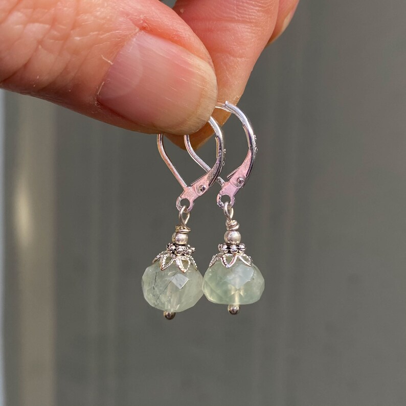 May include: A pair of silver leverback earrings with faceted, pale green gemstone drops. The earrings have decorative silver caps and small silver beads at the bottom of the gemstone drops. The earrings are held up by a hand.