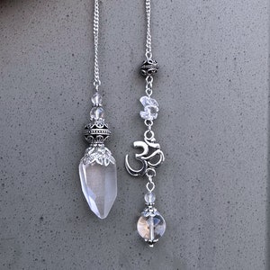 K&ouml;nnte beinhalten: Zwei silberfarbene Pendel mit klaren Kristallen und dekorativen Perlen. Eines hat einen klaren Quarz-Kristallpunkt und Zierarbeiten aus Metall. Das andere hat ein Om-Symbol und eine klare Kristallkugel.