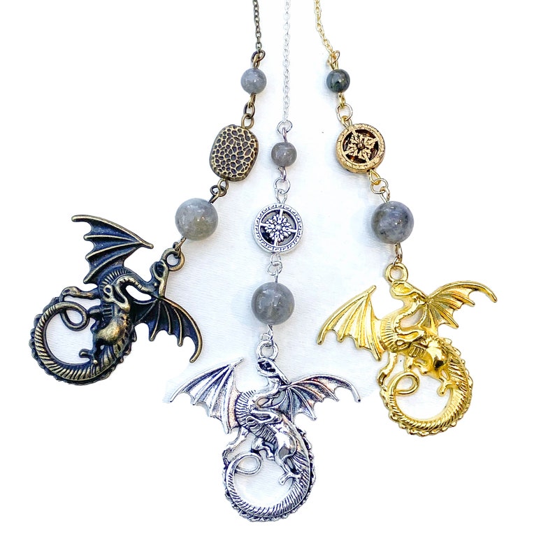 K&ouml;nnte beinhalten: Drei Drachenanh&auml;nger in Bronze, Silber und Gold. Jeder Anh&auml;nger zeigt ein detailliertes Drachendesign mit ausgebreiteten Fl&uuml;geln. Die Anh&auml;nger sind an Ketten aufgeh&auml;ngt, verziert mit grauen Edelsteinperlen und dekorativen Elementen.
