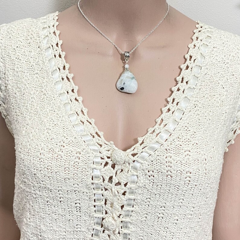 May include: A silver necklace with a teardrop-shaped pendant. The pendant is a white stone with black and gray inclusions. The necklace is displayed on a cream-colored crocheted top.