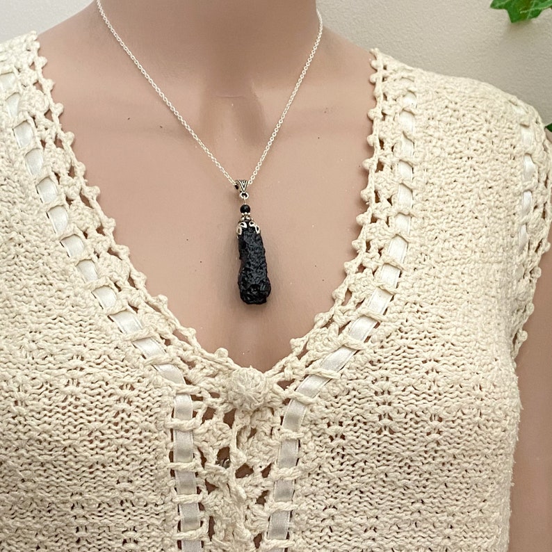 May include: A silver chain necklace with a dark, textured pendant. The pendant is a dark, irregular shape, possibly a stone or mineral. The necklace is worn over a cream-colored, open-weave top.