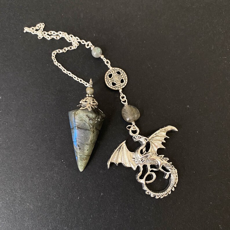 K&ouml;nnte beinhalten: Ein silberfarbenes Pendel mit einem facettierten, grau-gr&uuml;nen Stein und einem Drachenanh&auml;nger. Das Pendel h&auml;ngt an einer silbernen Kette mit grauen Steinperlen und einem dekorativen kreisf&ouml;rmigen Element. Der Drachenanh&auml;nger ist detailliert.