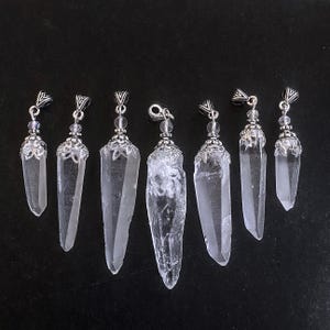 May include: Seven clear quartz crystal pendants with silver-colored metal caps and hanging loops. The crystals vary in size and shape, with some having a rough, natural texture. The background is a solid black.