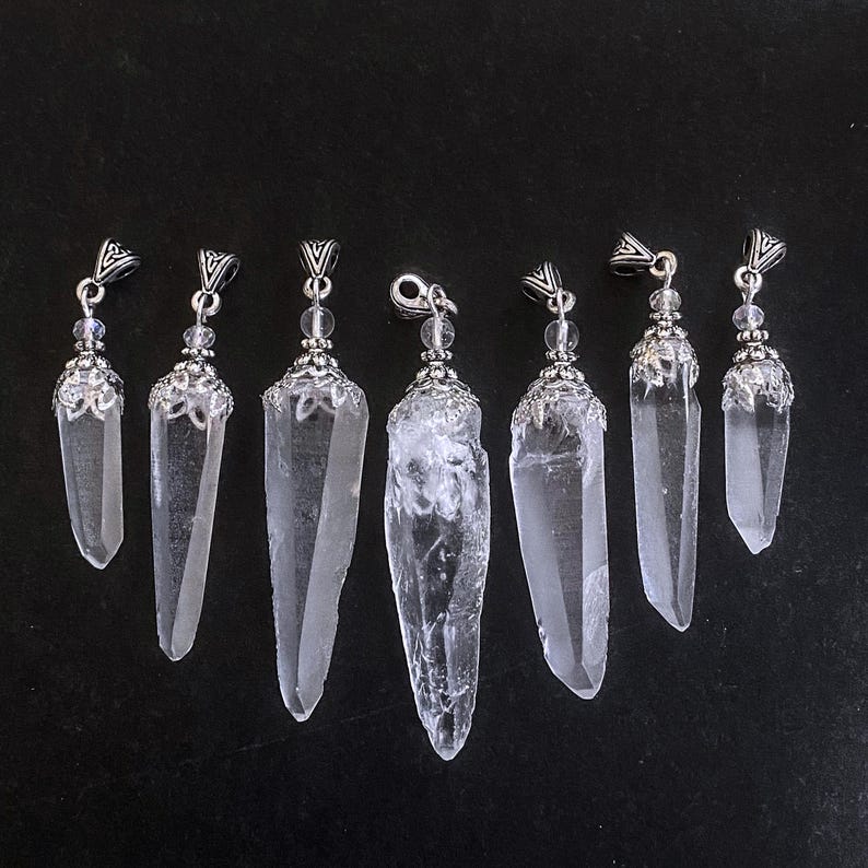 May include: Seven clear quartz crystal pendants with silver-colored metal caps and hanging loops. The crystals vary in size and shape, with some having a rough, natural texture. The background is a solid black.