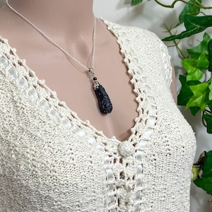 May include: A silver necklace with a dark, textured pendant. The pendant is a dark, elongated shape with a silver cap and a small red bead. The necklace is displayed on a cream-colored, crocheted top.