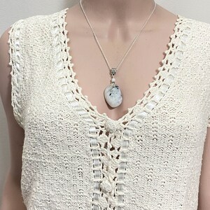 May include: A silver necklace with a large, irregular-shaped pendant. The pendant is a mix of white and gray, with a small silver accent at the top. The necklace is displayed on a cream-colored, crocheted top.