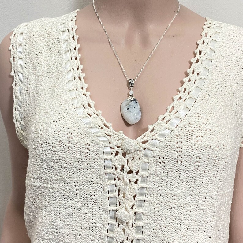 May include: A silver necklace with a large, irregular-shaped pendant. The pendant is a mix of white and gray, with a small silver accent at the top. The necklace is displayed on a cream-colored, crocheted top.