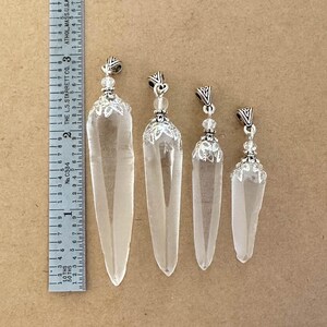 May include: Four clear quartz crystal pendants of varying sizes, each with a silver-colored metal cap and decorative beadwork. The largest pendant is approximately 3 inches long. The pendants are displayed on a neutral-toned surface.