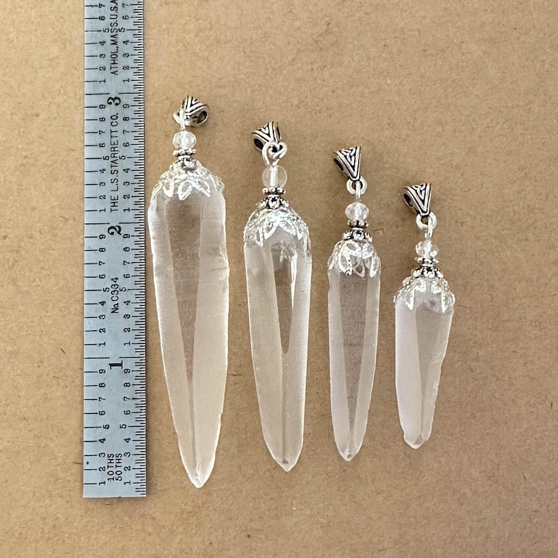 May include: Four clear quartz crystal pendants of varying sizes, each with a silver-colored metal cap and decorative beadwork. The largest pendant is approximately 3 inches long. The pendants are displayed on a neutral-toned surface.
