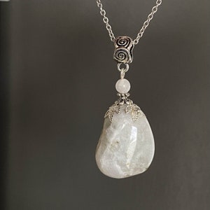 May include: A silver-toned necklace featuring a large, smooth, pear-shaped, off-white stone pendant. The pendant is accented with a small, round, light pink bead and ornate silver metalwork. The chain has a spiral design at the top.