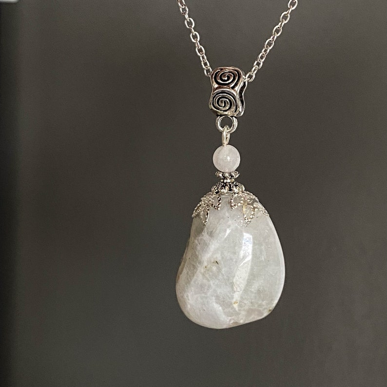 May include: A silver-toned necklace featuring a large, smooth, pear-shaped, off-white stone pendant. The pendant is accented with a small, round, light pink bead and ornate silver metalwork. The chain has a spiral design at the top.