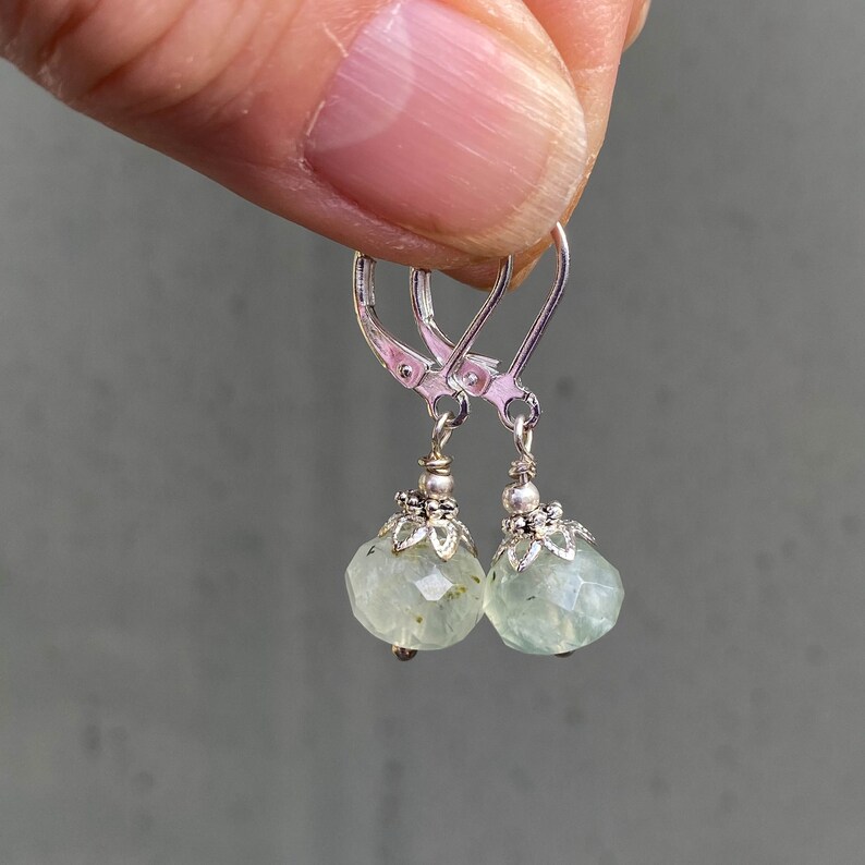 May include: A pair of silver leverback earrings with faceted, pale green gemstone drops. The gemstones have a round shape and are topped with decorative silver caps. The earrings are displayed against a blurred gray background.