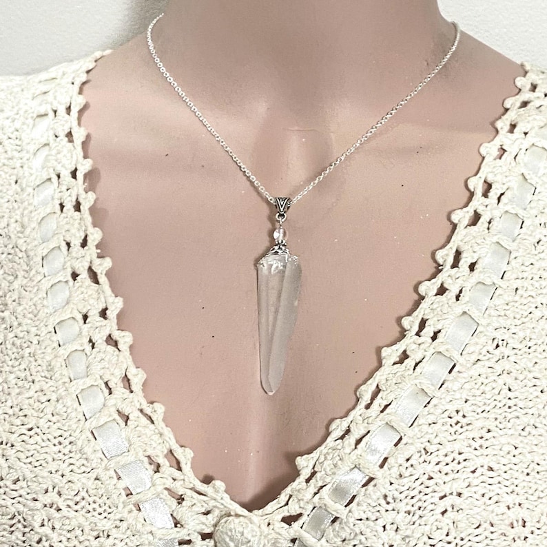 May include: A clear quartz crystal pendant necklace with a silver chain. The crystal is elongated and pointed, suspended from a decorative silver bail. The necklace is displayed against a cream-colored, crocheted top.