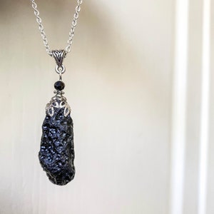 May include: A silver-toned necklace featuring a dark, textured, teardrop-shaped pendant. The pendant is suspended from a delicate chain and adorned with a small black bead and decorative metalwork. The necklace is displayed against a soft, neutral background.