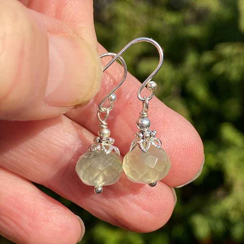 May include: A pair of silver dangle earrings. Each earring features a faceted, pale green gemstone pendant suspended from a decorative silver cap and a silver hook. The earrings are held up by a person's hand.