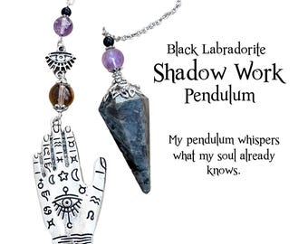 May include: A black labradorite pendulum with a silver chain and decorative beads. The pendulum is labeled "Black Labradorite Shadow Work Pendulum" with the text "My pendulum whispers what my soul already knows."