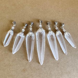 May include: Seven clear quartz crystal pendants with silver-colored metal caps and decorative accents. Each crystal is elongated and pointed, with a polished, transparent appearance. The pendants are arranged on a light brown surface.