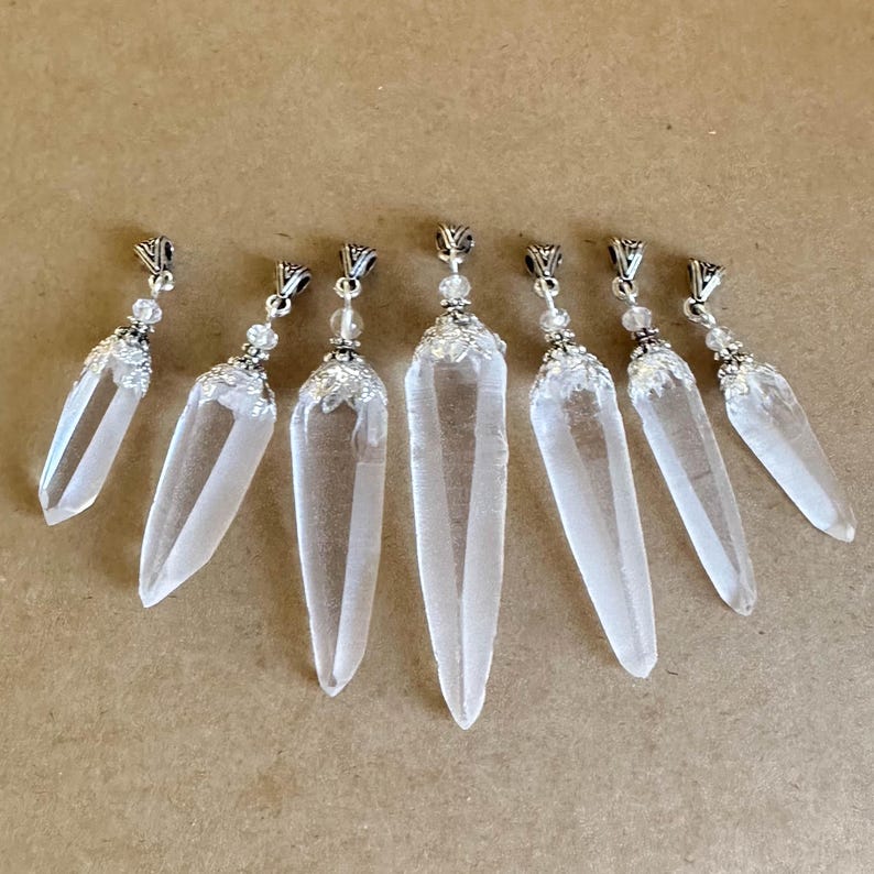 May include: Seven clear quartz crystal pendants with silver-colored metal caps and decorative accents. Each crystal is elongated and pointed, with a polished, transparent appearance. The pendants are arranged on a light brown surface.