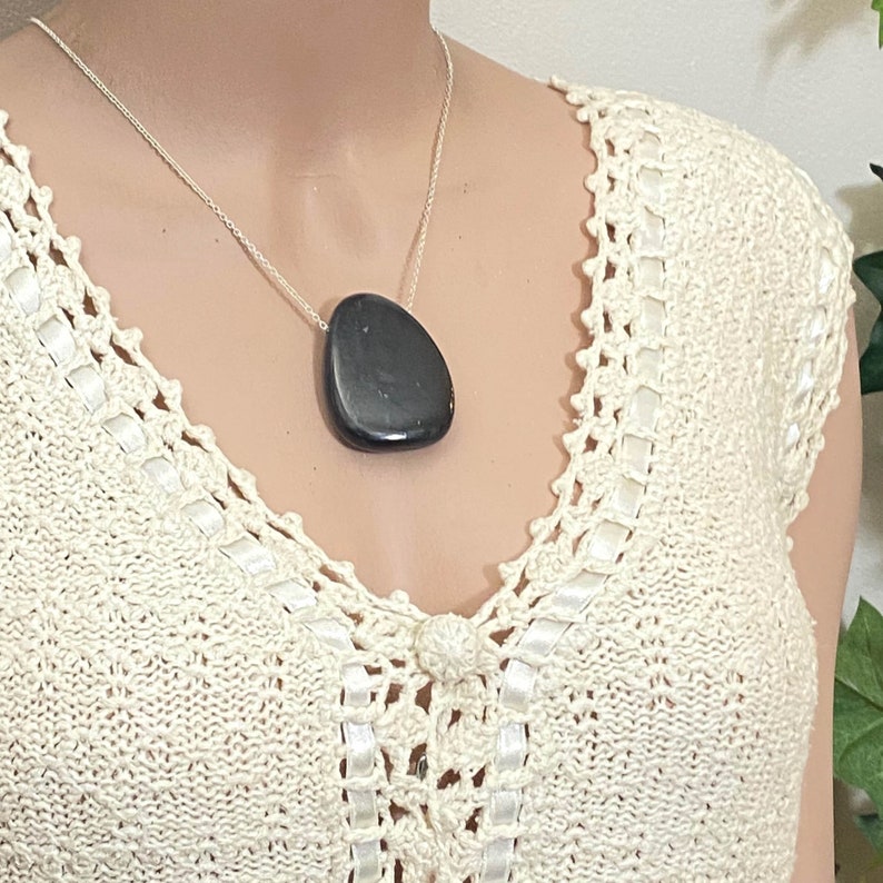 May include: A silver chain necklace with a large, dark gray, teardrop-shaped pendant. The pendant is smooth and polished. The necklace is displayed on a person wearing a cream-colored, crocheted top with white ribbon accents.