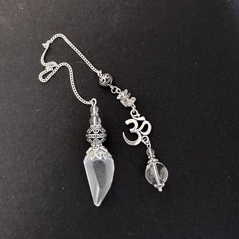 K&ouml;nnte beinhalten: Ein silberfarbenes Pendel mit einer Spitze aus klarem Kristall und dekorativen Metallperlen. Die Kette ist mit einem Anh&auml;nger mit dem Om-Symbol und klaren Kristallperlen verbunden. Der Hintergrund ist schwarz.