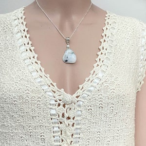 May include: A silver chain necklace with a pendant featuring a white and gray stone. The stone is teardrop-shaped and has a polished finish. The necklace is displayed on a cream-colored crocheted top.