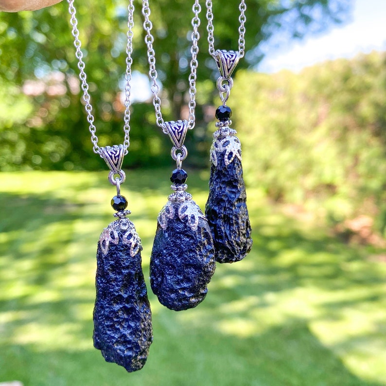 May include: Three black, textured pendant necklaces with silver-colored chains and decorative caps. Each pendant has a unique, elongated shape and is accented with a small black bead. The background is a blurred green, suggesting an outdoor setting.