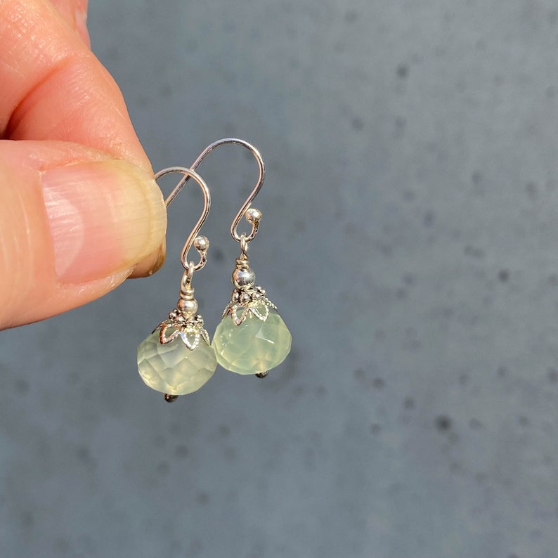 May include: A pair of silver earrings with faceted, pale green teardrop-shaped stones. The earrings have a decorative silver cap and a simple hook closure. The earrings are held up against a gray background.