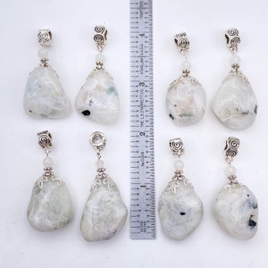May include: Eight polished, teardrop-shaped moonstone pendants with silver-colored metal accents. Each pendant is approximately 1.5 inches long. The stones are white with subtle blue and gray inclusions. A ruler is placed next to the pendants.