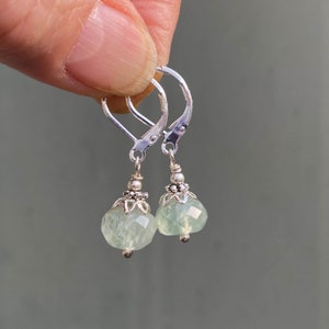 May include: A pair of leverback earrings with faceted, pale green gemstone drops. The earrings have silver-colored metal components, including the leverback closures, decorative caps, and small beads. The gemstones are a soft, translucent green color.