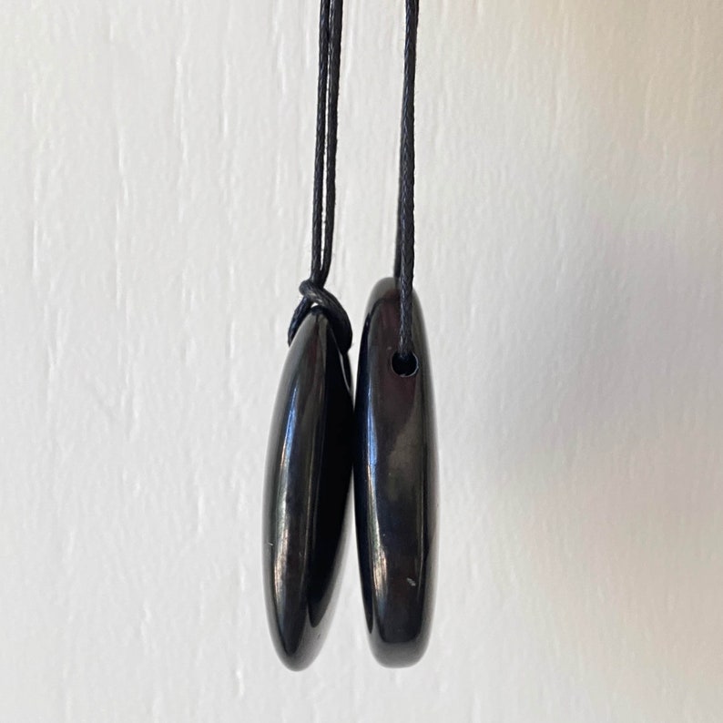 May include: Two black, teardrop-shaped pendants hanging from black cords. The pendants have a smooth, glossy finish and are suspended against a white background. The cords are knotted together at the top.