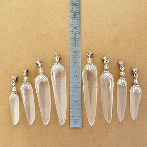 May include: Clear quartz crystal pendants of varying sizes, ranging from approximately 1 to 3 inches in length, with silver-colored metal caps and decorative accents. The pendants are displayed on a neutral-toned surface.