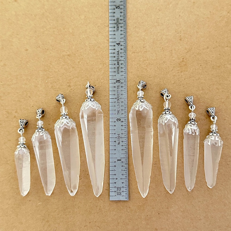 May include: Clear quartz crystal pendants of varying sizes, ranging from approximately 1 to 3 inches in length, with silver-colored metal caps and decorative accents. The pendants are displayed on a neutral-toned surface.