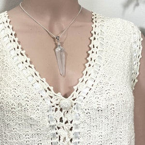 May include: A silver necklace with a clear, pointed crystal pendant. The crystal is suspended from a delicate silver chain. The necklace is displayed against a cream-colored, crocheted top with a V-neck design.