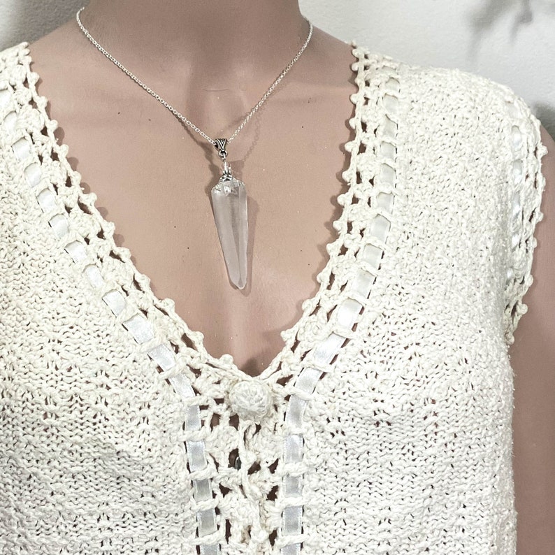 May include: A silver necklace with a clear, pointed crystal pendant. The crystal is suspended from a delicate silver chain. The necklace is displayed against a cream-colored, crocheted top with a V-neck design.