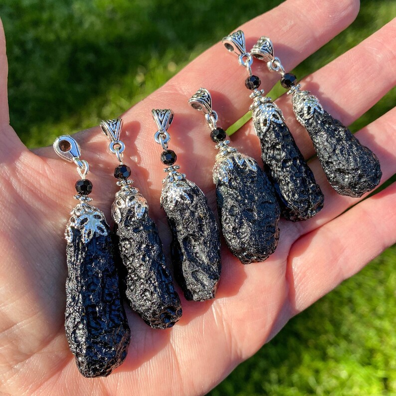 May include: Six black pendants with textured surfaces and silver-colored metal accents are displayed. Each pendant has a unique shape and is adorned with a small black bead. The pendants are held in a hand against a backdrop of green grass.