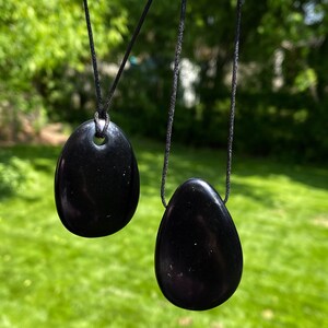 May include: Two black, oval-shaped pendants hanging from black cords. The pendants have a smooth, polished surface and a hole at the top for the cord. The background is a blurred view of green grass and trees.