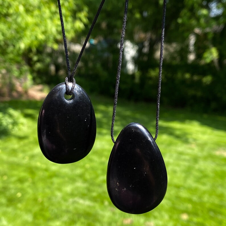 May include: Two black, oval-shaped pendants hanging from black cords. The pendants have a smooth, polished surface and a hole at the top for the cord. The background is a blurred view of green grass and trees.