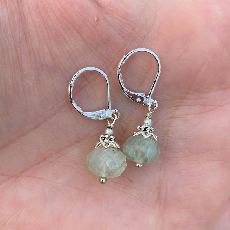 May include: A pair of silver leverback earrings with faceted, pale green gemstone drops. The earrings feature intricate silver beadwork and a small silver bead at the base of each drop. The earrings are displayed against a light background.
