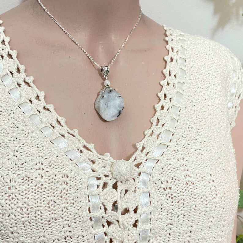 May include: A silver necklace with a large, irregular-shaped, white and gray moonstone pendant. The pendant hangs from a delicate silver chain. The necklace is displayed on a cream-colored, crocheted top.