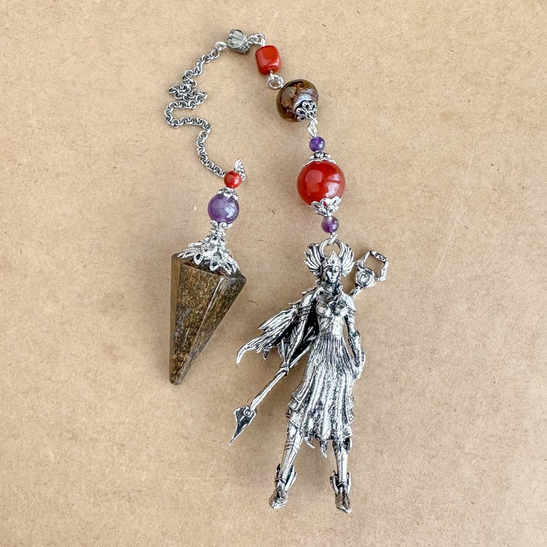 May include: A divination pendulum with a silver chain and decorative beads. The pendulum has a faceted, brown stone, a silver figurine, and beads in red, purple, and brown. The figurine is of a woman with a spear and a shield.