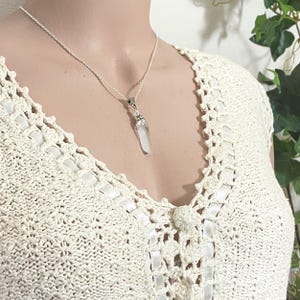 May include: A silver necklace with a clear quartz crystal pendant. The crystal is elongated and faceted. The necklace is displayed on a cream-colored crocheted top.