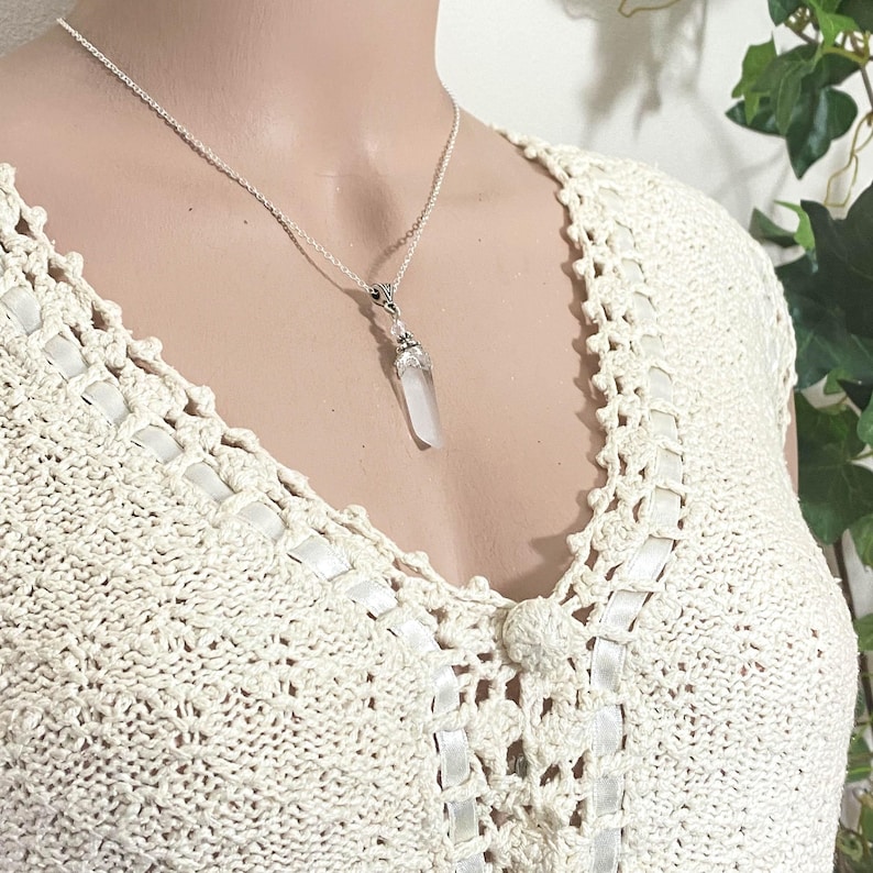 May include: A silver necklace with a clear quartz crystal pendant. The crystal is elongated and faceted. The necklace is displayed on a cream-colored crocheted top.