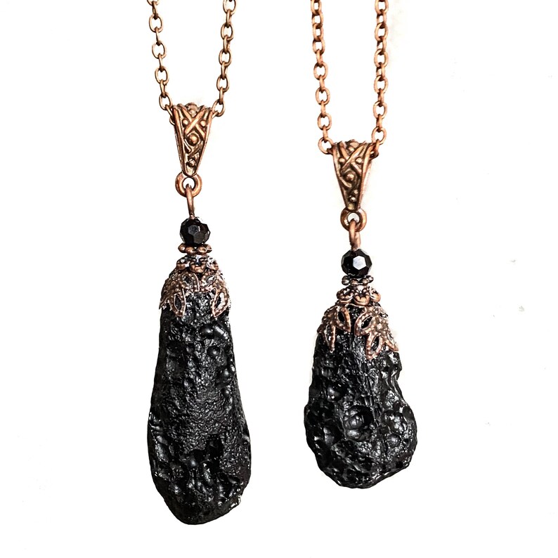 May include: Two pendant necklaces. Each features a dark, textured, teardrop-shaped stone suspended from a copper-colored chain. The pendants have ornate copper-colored caps and a black bead. The chains are delicate and the background is white.
