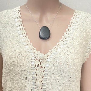 May include: A silver necklace featuring a large, smooth, dark gray stone pendant. The pendant is teardrop-shaped. The necklace is displayed on a cream-colored, open-weave top with a V-neckline and decorative trim.