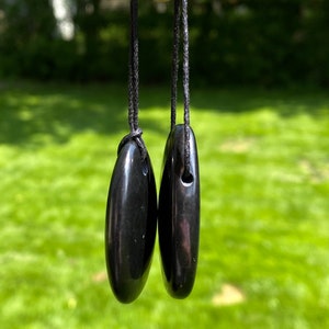 May include: Two polished black oval pendants hanging from black cords. The pendants are smooth and glossy, reflecting the light. The background is a blurred green lawn.