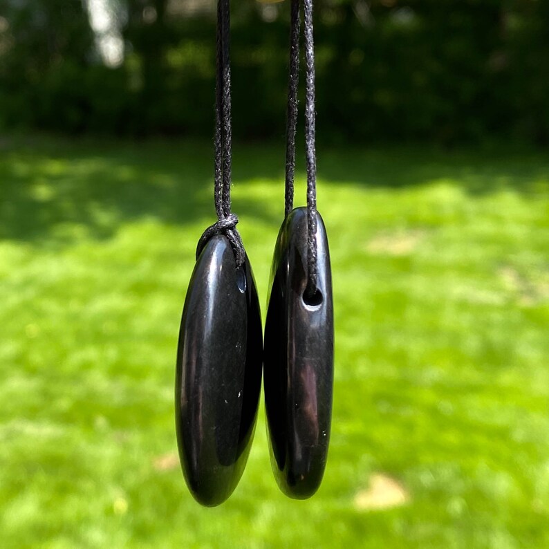May include: Two polished black oval pendants hanging from black cords. The pendants are smooth and glossy, reflecting the light. The background is a blurred green lawn.